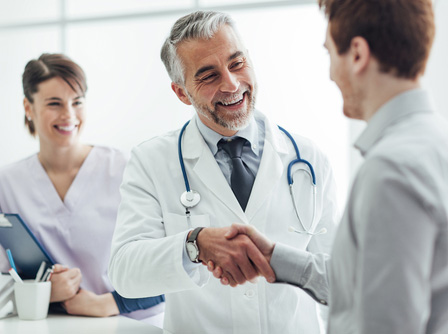 Picture of a smiling Physician shaking a mans hand. There is a Nurse behind him holding a clipboard and smiling.