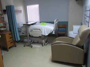 Picture of a hospital patients room. There is a reclining chair, mini fridge, microwave, small chair, bedside table, bedside table tray, swing bed, privacy curtain, window, and a dry eraser board.