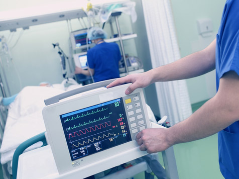 Picture of a heart monitor machine in pre-op at the foot of the bed. There is a female Nurse in the background putting patient info into a computer and patient lying down next to the Nurse in a swing bed.