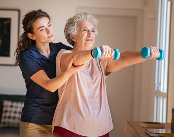 Picture of a female Physical Therapist standing behind a female patient and she has her hands under her elbows while she supports her holding weights straight out in front of her.