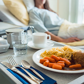 Picture of a female patient sitting up in a swing bed looking over at a bedside table tray that has fried chicken, carrots, and noodles on a plate, silverware, coffee cup, and glass cup.