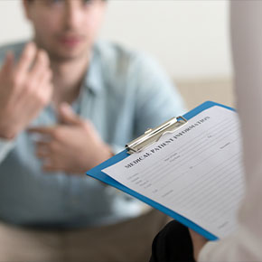 Picture of a male patient sitting down on a couch looking up at a female  Physician who is sitting in front of him holding a clipboard with Medical Forms on it.