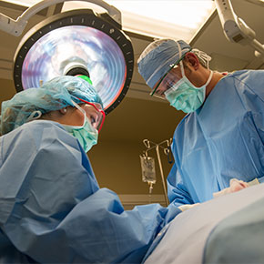 Picture of two surgeons (male and female) from a ground shot looking up. They are wearing surgical covers, mask, surgical glasses, surgical head cap, and gloves and they are standing over a patient.