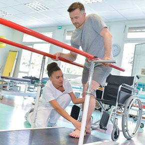 Picture of a female Nurse helping assist a male patient. He is standing up from his wheel chair and grabbing hand rails for support to start walking. The female Physical Therapist is pushing his foot down to the ground and holding his knee straight.